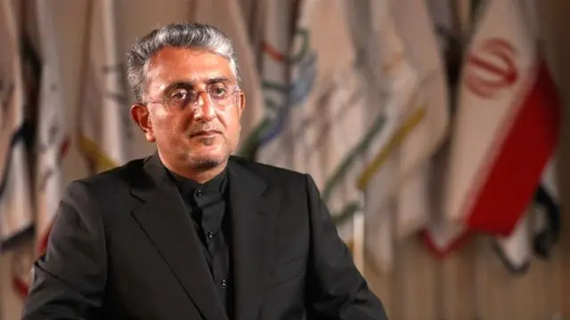 رجل يرتدي قميصاً وسترة باللون الأسود وينظر إلى الكاميرا، جالساً وخلفه أعلام، بينها العلم الإيراني.