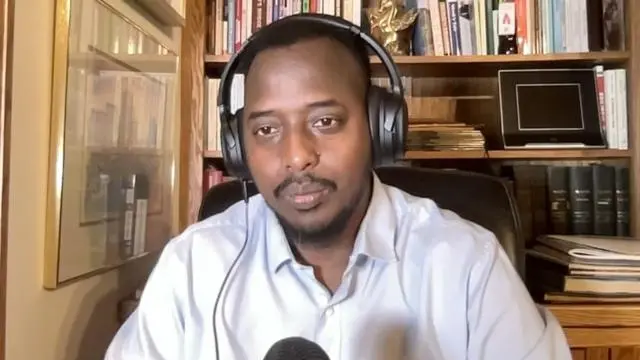 رجل في مكتب يرتدي سماعات رأس تغطي الأذنين وقميصاً أبيض، ويتحدث في ميكروفون.