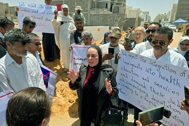 سيدة تخاطب مجموعة من المتظاهرين الذين حملوا لافتات كتب على إحداها:
"We reject the integration of immigrants into health education and services and demand their ... compulsory deportation"